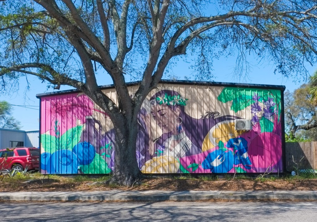 a tree in front of a mural on the side of a building, of a woman surrounded by fresh fruit, lemon, watermelon, blueberries, mango