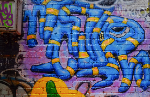 painted street art on a brick wall, blue and yellow striped snake like creature with one big eye and a small mouth with yellow teeth, looks to be smiling
