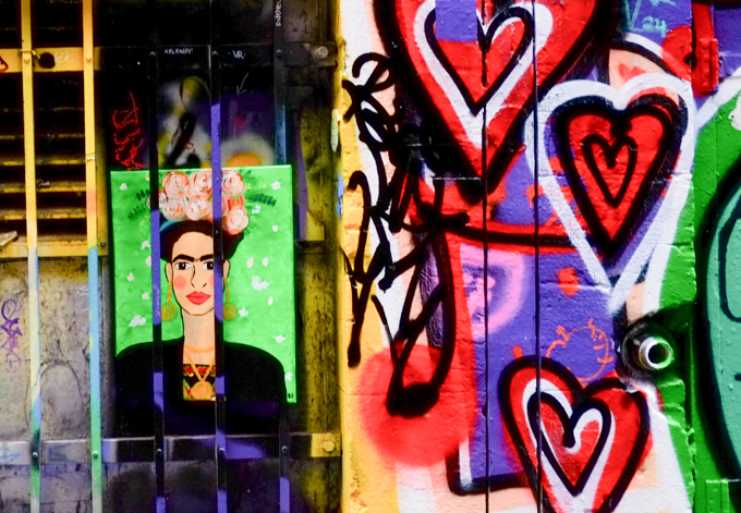 Painting of Frida Kahlo, graffiti, behind bars, in an alley, with many hearts painted beside it