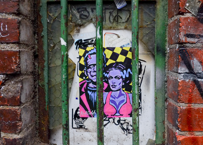 graffiti poster by Ryan Callisto, a purple Frankenstein, along with a purple bride of Frankenstein, on a wall behind green metal bars, in an alley