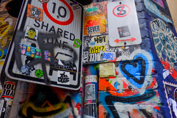 In an alley, wall and shared street zone  signs are covered with stickers and spray paint graffiti