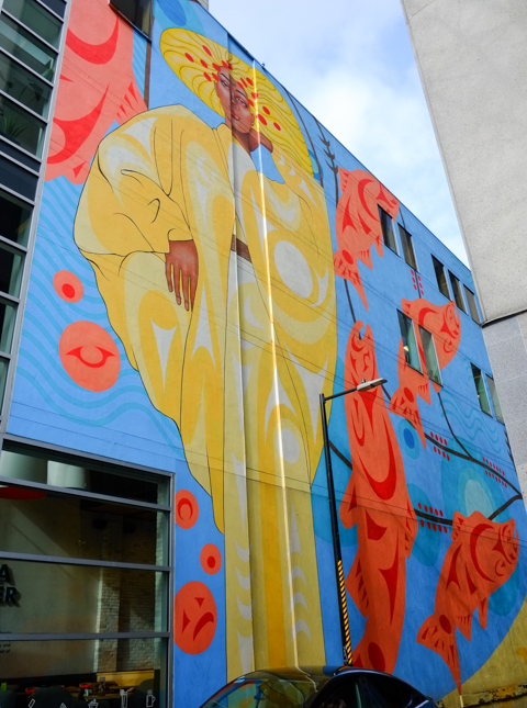 large mural of woman in long yellow outfit, orange salmon fish around her