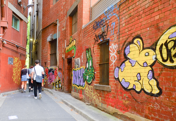people walking through a narrow alley from Langs Lane, red brick buildings beside, with graffiti on the walls 