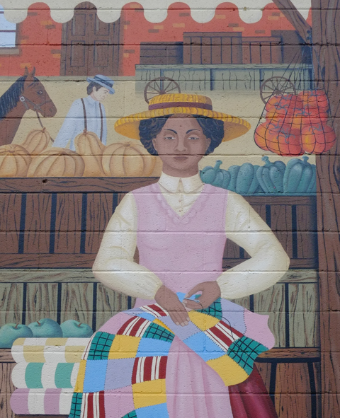 part of a history of chatham mural, market scene, woman in hat is sewing a quilt as she sits in a store