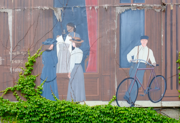 history of Chatham mural, older, peeling and faded a bit, a line of businesses, two women looking in the window of Lyons tailoring where a woman is adjusting a dress on a headless mannequin.  A man about to get on a bike is also in the picture