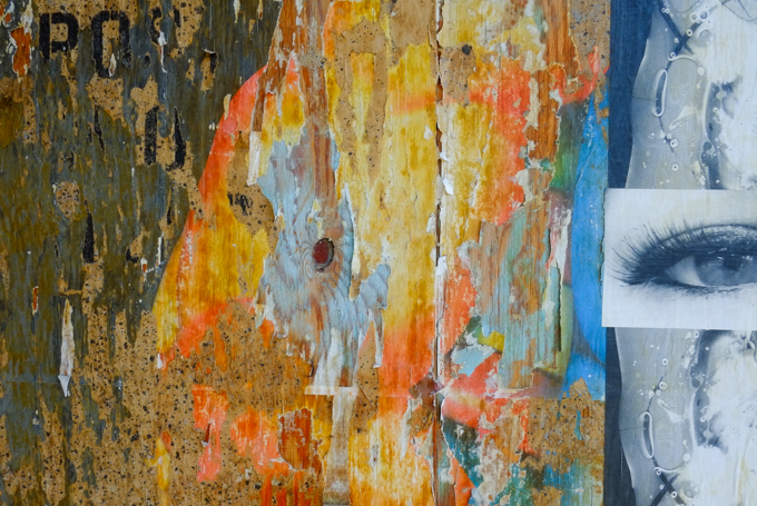 an eye, and a sliver of the edge of a paper paste up piece, beside a section of plywood wall with multiple bits of old paper from graffiti, and some paint