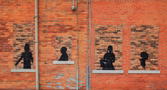 on a brick wall, onld windows have been bricked over and in each of the 4 windows is a black silhouette that looks like a famous film character, Edgar Allan Poe, Dorothy and Toto from the Wizard of Oz, Willy Wonka, and the little spymaster woman Edna in the "The Incredibles' movies