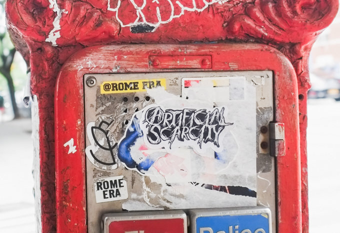red emergency call box in new york city with sticker graffiti on it including one that says artificial scarcity