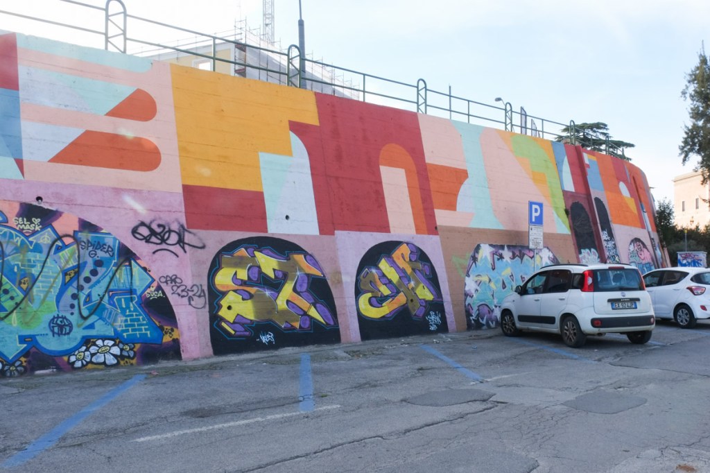 curved wall, exterior, painted, top level in shapes, by Eltono, bottom part in arch shapes with each arch filled in by a different artist
