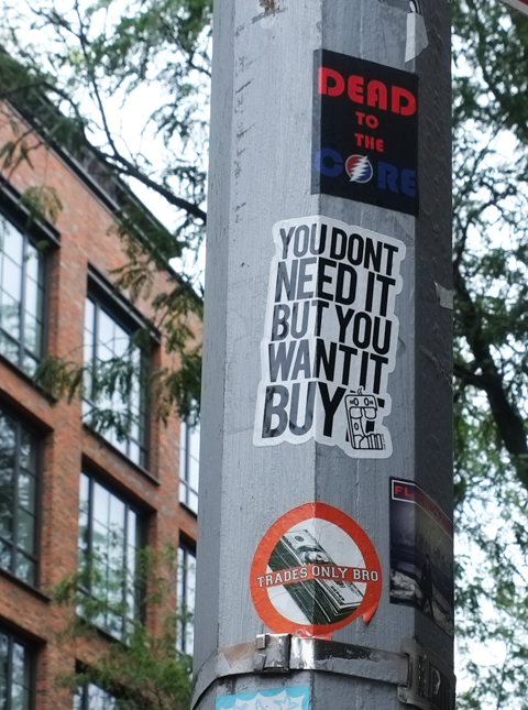 graffiti sign on a pole with words that say You don't need it but you want to buy it