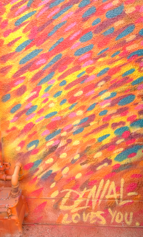 part of a larger street art painting, dabs of color in orange, red, yellow, and blue, in a diagonal pattern, with text at bottom that says Denial loves you 