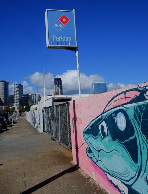 old sign for parking for Dominos Pizza stands above a long concrete block fence that has been painted with murals. at one end is a big blue fish head on pink background
