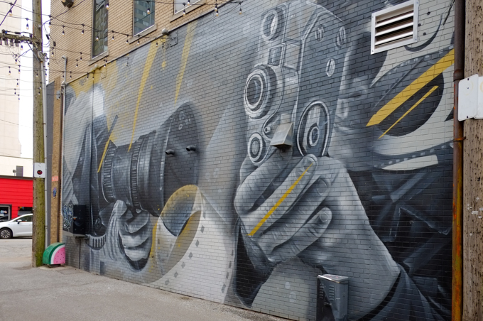 large mural in an alley by Derkz, shades of grey, very realistic, people with movie cameras, one modern, one slightly older, hand held cameras, 