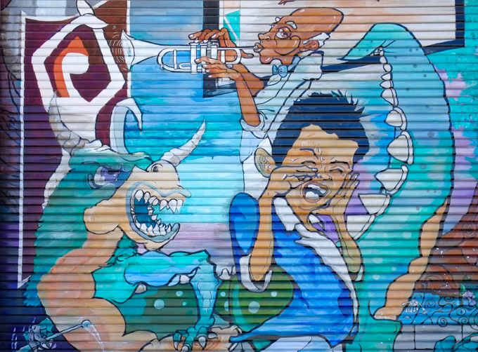 part of a street art mural, a young man with eyes closed, an older man playing the trumpet, a dragon with open mouth, horns on head, and a long spiky tail.