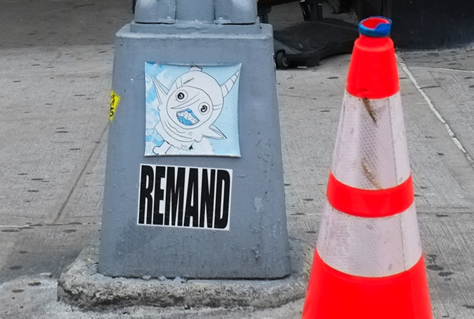 slap graffiti on a grey pole, one says remand in large black letters, the other is a drawing of a small creature with large head, big mouth, 