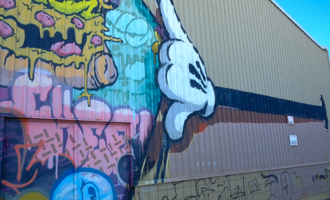 part of a street art mural, a very long mickey mouse arm (skinny, black, and with a white glove on the fat hand) reaches out of a hole in the wall and pushes aside the start of a painting