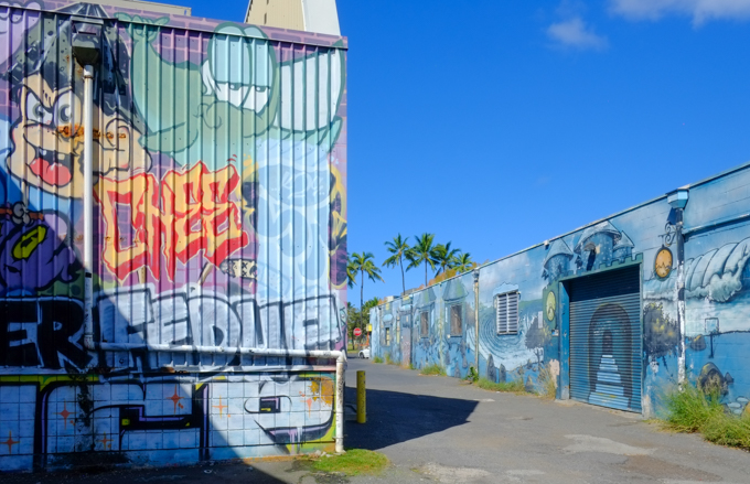 looking down an alley between two buildings, street art on both sides. on the left is an orange face, on the right is a blue mural by katch 1 that has a japanese theme