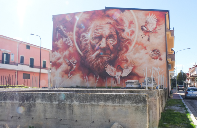 in a residential area of stornara, side of apartment building covered by mural by aero, large mural in shades of reds and orange, a face made of two elderly halves, on side male and the other side female, with 5 birds flying around