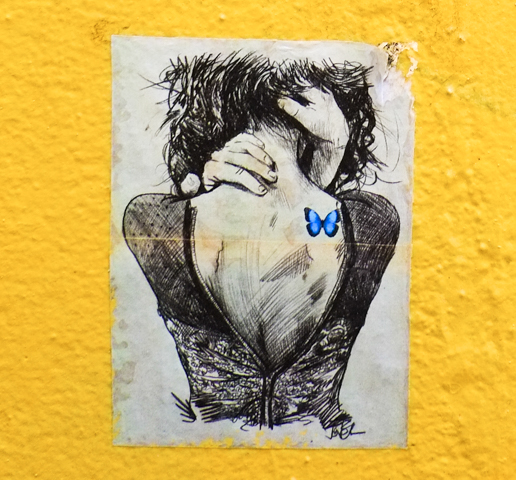 small paste up on a yellow wall in an alley, black and white drawing of a woman's back, long hair, blue butterfly tattoo on one shoulder