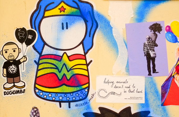 stickers and slaps on a door, one is DJ Gumba with two black balloons, one is a wonder woman figure by little meh, and one is a man in a plaid shirt carrying a very large carrot also a sticker with just text that reads helping animals doesn't need to be that hard 