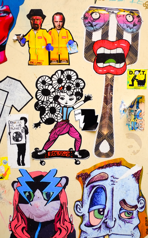 many stickers and paste ups on a door, there is no planet B, a drecks woman on a skateboard, two breaking bad lego figures in yellow, a spatula turned into a puppet head with big red lips, 