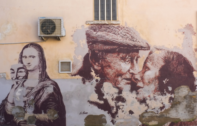 2 pieces of street art, one is Mona Lisa holding up an anonymous mask and the other is an elderly couple hugging. 