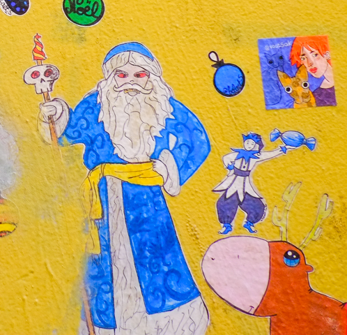 small Christmas themed paste ups on a yellow wall in Melbourne, Baptist Place, Father Christmas with long white hair and beard, in a blue floor length robe, holding a staff with a skull on it, also a wall lilo figure holding a candy wrapped in blue wrapper