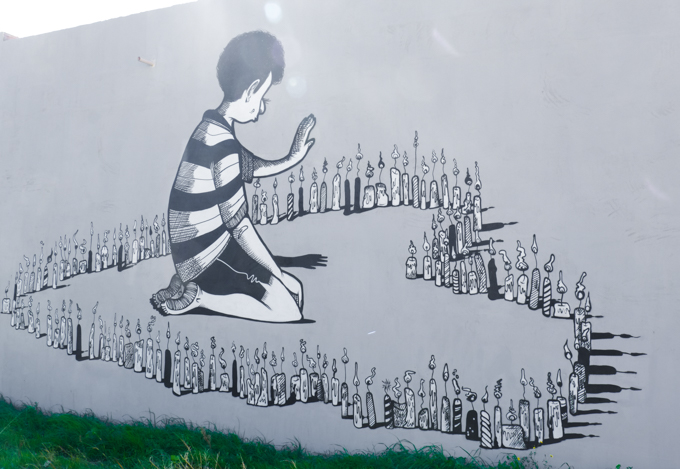 in shades of grey, a mural by Alex Senna of a young boy kneeling by a large heart shape on the ground made of lit candles