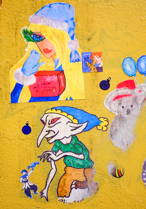 small Christmas themed paste ups on a yellow wall in Melbourne, Baptist Place, a koala in a red hat, a long nosed elf accepting a candy from a wall lilo figure, and a woman wearing fur cuffed mitts