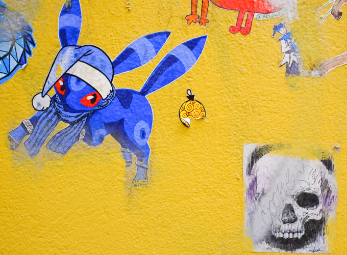 graffiti on a yellow wall, a blue pokeman creature with blue cap with white pompom and a blue scarf, also a black and white drawing of a skull by Loui Jover 