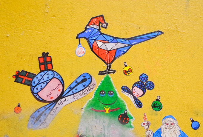 small Christmas themed paste ups on a yellow wall in Melbourne, Baptist Place, a smiling green Christmas tree, with a red, white and blue bird on top, two sleeping girls by balaets, one with wrapped gifts on her head, sleeping and dreaming, bird has Christmas ornament in its mouth