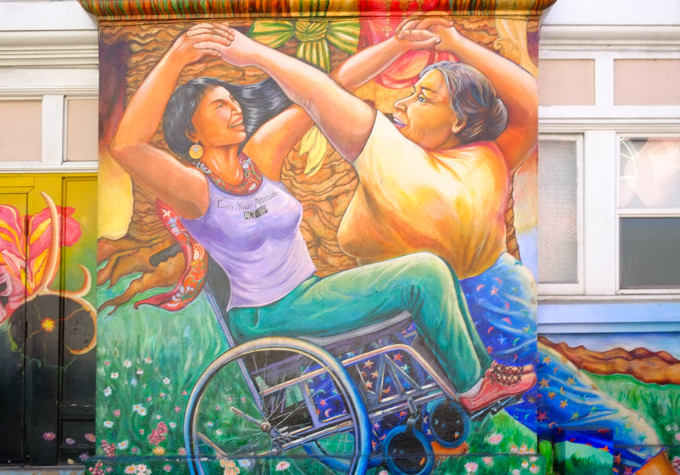 part of maestrapeace, a mural on the womens building, women holding hands above their heads and dancing, one of whom is in a wheelchair
