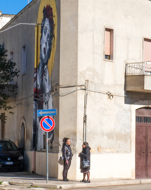 two women standing on a street corner talking to each other, one of the buildings beside them has a street art portrait