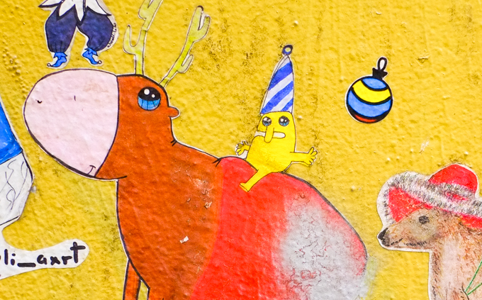 small Christmas themed paste ups on a yellow wall in Melbourne, Baptist Place, a silly little yellow character wearing a tall blue and white striped hat and sitting on the back of a brown animal with yellow antlers