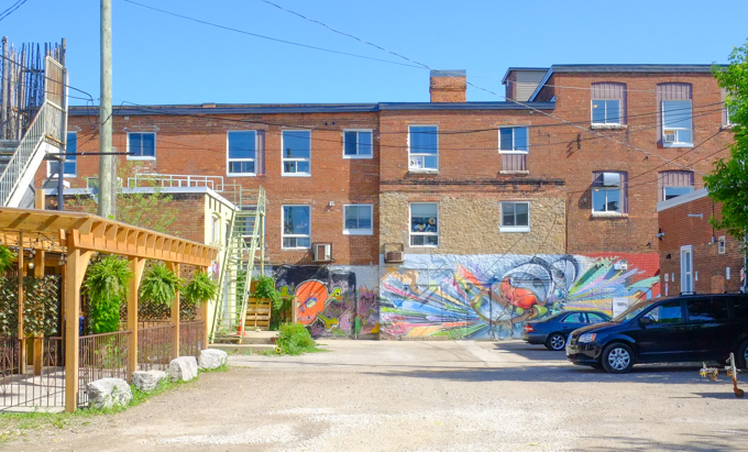 parking lot and alley behind shops in Own Sound, three storey brick buildings with street art murals along the bottom third.