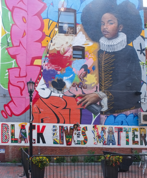 mural by Jeff Henriquez in Springfield Mass, young black man dressed in Dutch renaissance clothing including white collar and large black hat, with a can of spray paint and a painters palette.  across the bottom are the words black lives matter