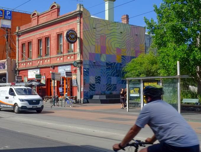 cyclist cycling past a street art mural on the side of an old two storey building on Sydney Road in Brunswick