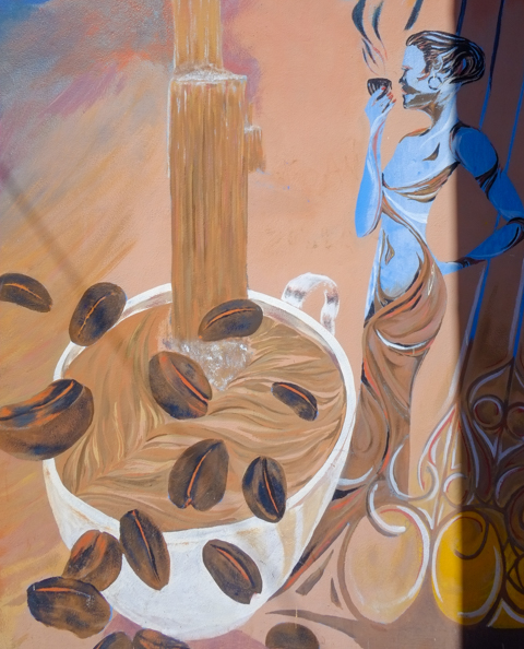 mural, large white coffee mug with coffee being poured into it, lots of coffee beans hopping around, a blue woman in long brown dress is holding a seraming cup of coffee