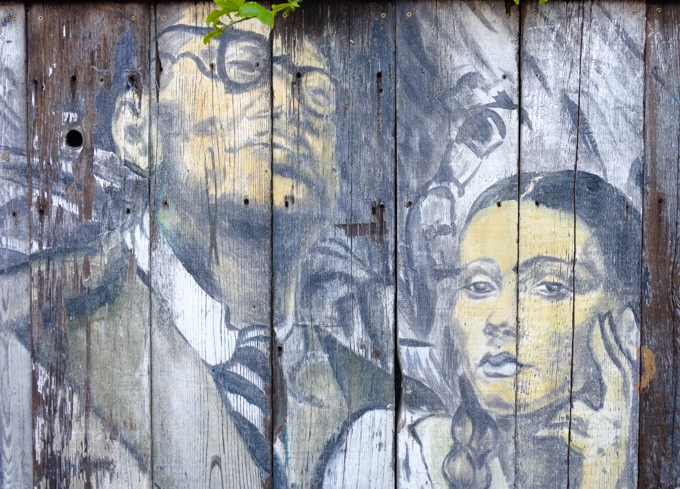 faded mural on wood fence, two people, man and woman, man wearing glasses, woman with long braids 