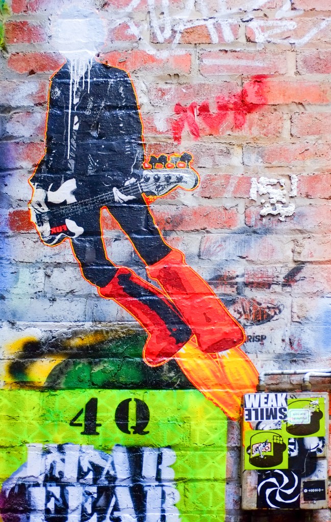 urban art in AC DC lane in Melbourne, headless (white paint sprayed over head) guitar player with big red boots that are also flaming rockets, stencil on brick wall