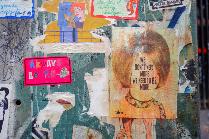 urban art in AC DC lane in Melbourne, paste up by Jover of woman's head but instead of facial features, has text that says we don't need more, we need to be more.   another sticker that says always learn 