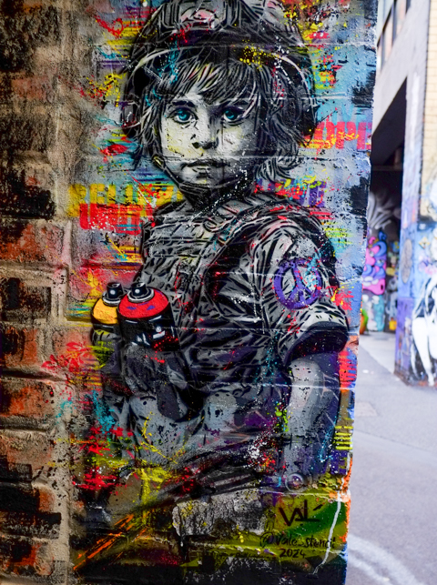 stencil of a blue eyed child in helmet and vest, spray paint cans on vest, peace symbol on sleeve of t shirt, 