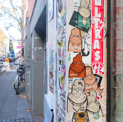 paste up graffiti on a wall, 7 cartoonish characters in a column, all in different colours, the word merde has been written over each one in white ink or white marker, 