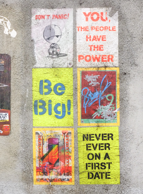 6 graffiti posters hung together on a concrete wall, most have text - don't panic, you the people have the power, be big, never ever on a first date, 