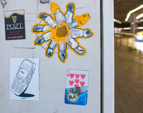 Three graffiti slaps, one is a line drawing of a mobile phone, another is a playing card thirteen of hearts, and the other is a pinwheel or gear cog made of robot arms or gloves in a circle