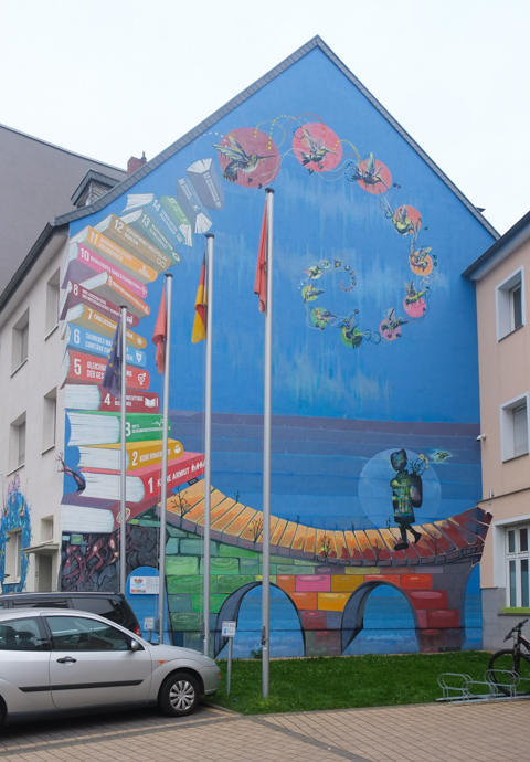 street art on Hochstadenring in Bonn Germany, mural on the side of building, blue sky and water background, books numbered 1 through 17, forming a curve that leads into a bridge that a child is walking on
