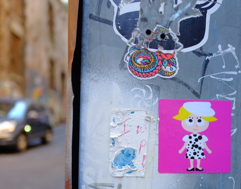 stickers and paste ups on a grey metal surface in Higson Lane, one is a girl with a black and white cow dress on a bright pink background
