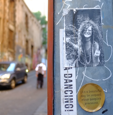 paper pasteups on a grey surface in Higson Lane, one is  a black and white photo of a young woman with long curly hair, she is smiling.  The other is gold disc with text that says It's a beautiful day to respect other people's pronouns. 