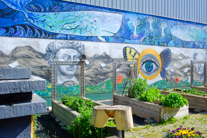 mural painting along one wall, exterior, outdoors, of the Grey Gallery Garden. Two large blue whales across the top, two male portraits below with a large eye with butterfly wings between the men