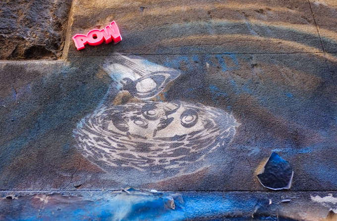 stencil street art of a surveillance camera by a bird's nest, also 3 D red letters spelling POW